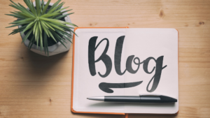 A paper with the word written Blog and a plant sitting on a desk. Used to describe why Blogging is important